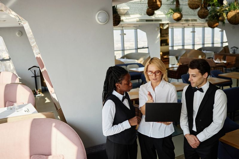 Middle aged Caucasian woman showing something on laptop to young adult Black woman and young adult Caucasian man in restaurant, all wearing uniforms and standing together