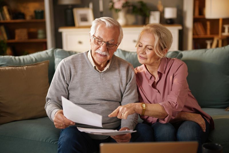 Elderly couple reviewing life insurance policy documents, planning their future finances together on the sofa