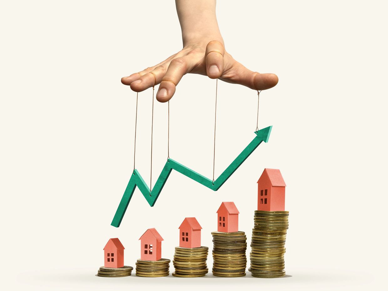 Hand manipulating a rising green arrow over houses on coin stacks.