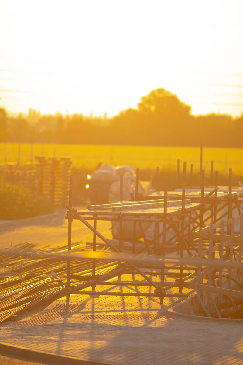 A stunning sunset paints vibrant colors over a busy construction site, with scaffolding in the foreground and a lush field behind, forming a beautiful scene
