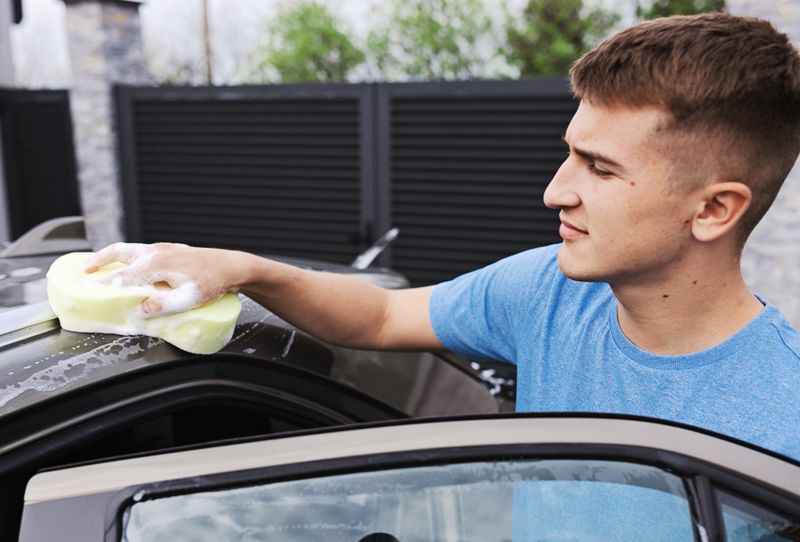 A man is thoroughly cleaning his car in an outdoor area using a sponge soaked with suds, ensuring it sparkles for better maintenance and appearance.