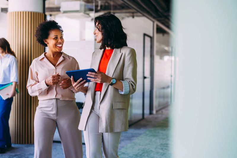 Two female professionals engaging in a discussion while standing in a contemporary open office setting, highlighting collaboration and productivity. Bright colors and natural interactions emphasize a positive workplace culture.
