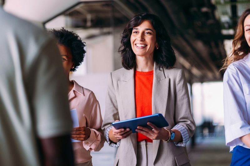 Smiling businesswoman holding a tablet, engaged in a collaborative workplace environment surrounded by colleagues in an open office setting.