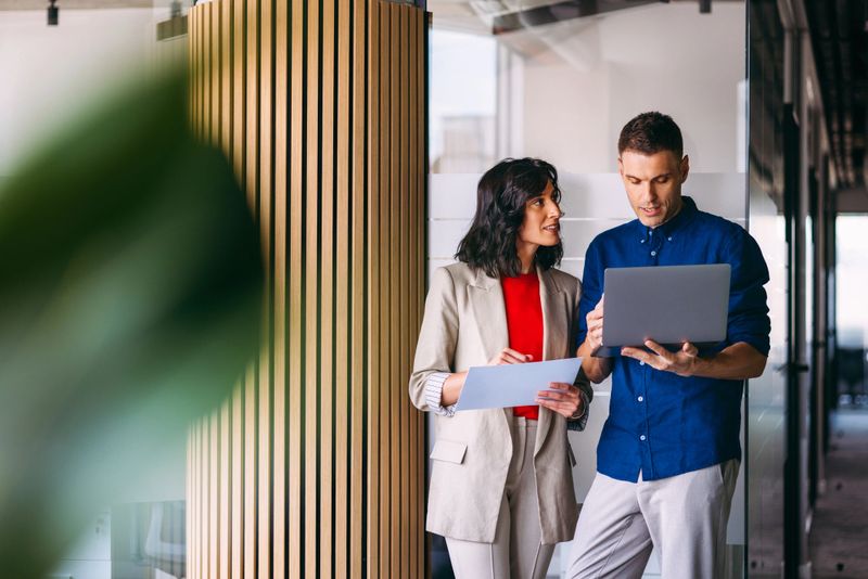 Two professionals discussing strategies using a laptop and documents, showcasing teamwork and productivity in a contemporary office setting.