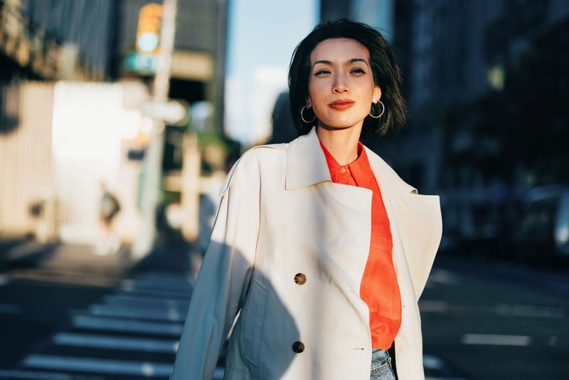 A stylish woman dressed in a white coat and red blouse smiles confidently as sunlight illuminates her surroundings on a city street, exuding elegance and sophistication.