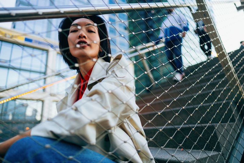Modern portrait of a woman seated behind a chain-link fence with contemporary architecture and urban elements, showcasing a casual and stylish fashion sense with a bold perspective.