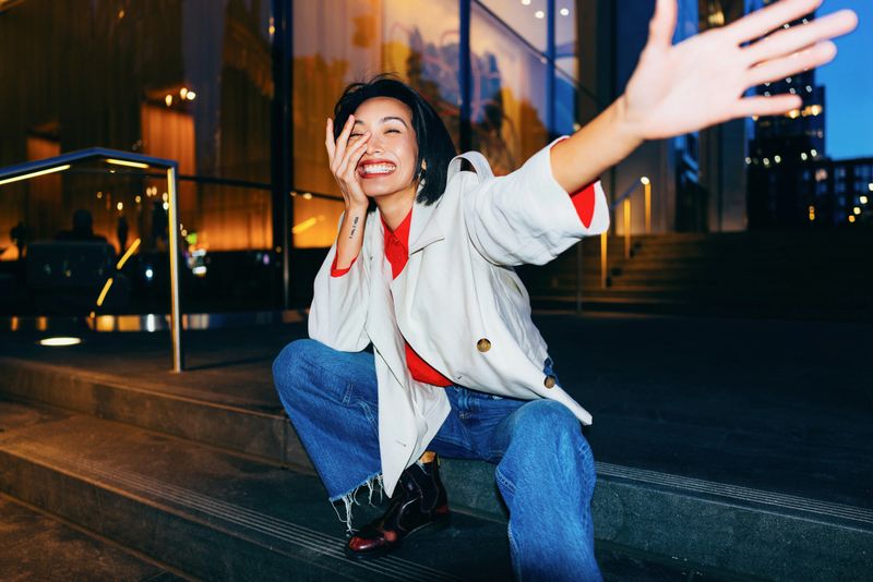 An expressive and joyful young woman sits on steps in an urban environment during nighttime, embracing positivity and excitement.
