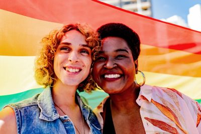 Two smiling people in front of a rainbow flag.