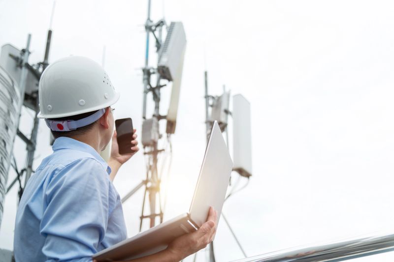 Engineer using mobile phone and laptop near 5G communications tower