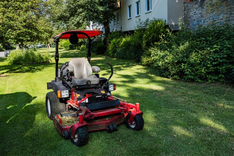 A riding mower is parked on freshly cut grass in a residential area. The green lawns and surrounding trees indicate a well-maintained outdoor space during daylight hours.