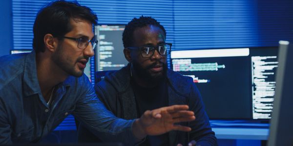 Two programmers intensely discussing code on multiple monitors in a dimly lit room.