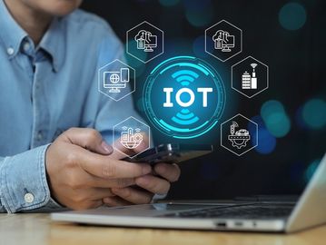 Person holding smartphone with IoT technology icons and laptop on table.