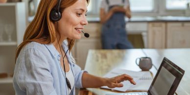 Woman with headset working on laptop at home, another person in background.