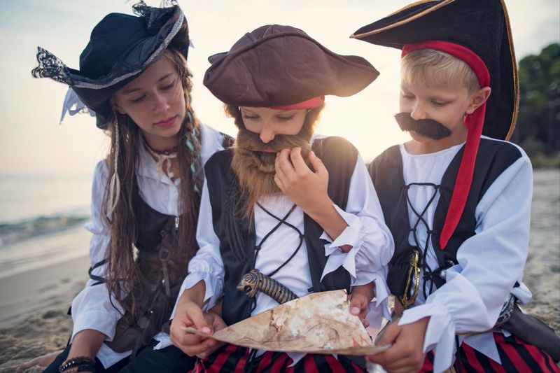 Three kids dressed in full pirate costumes are immersed in a playful scene on a sandy beach. Kids are kneeling on sand, reading directions from a treasure map.Sunny summer day.Shot with Nikon D850