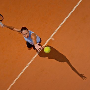 Tennis player reaching for a ball on a clay court from above.