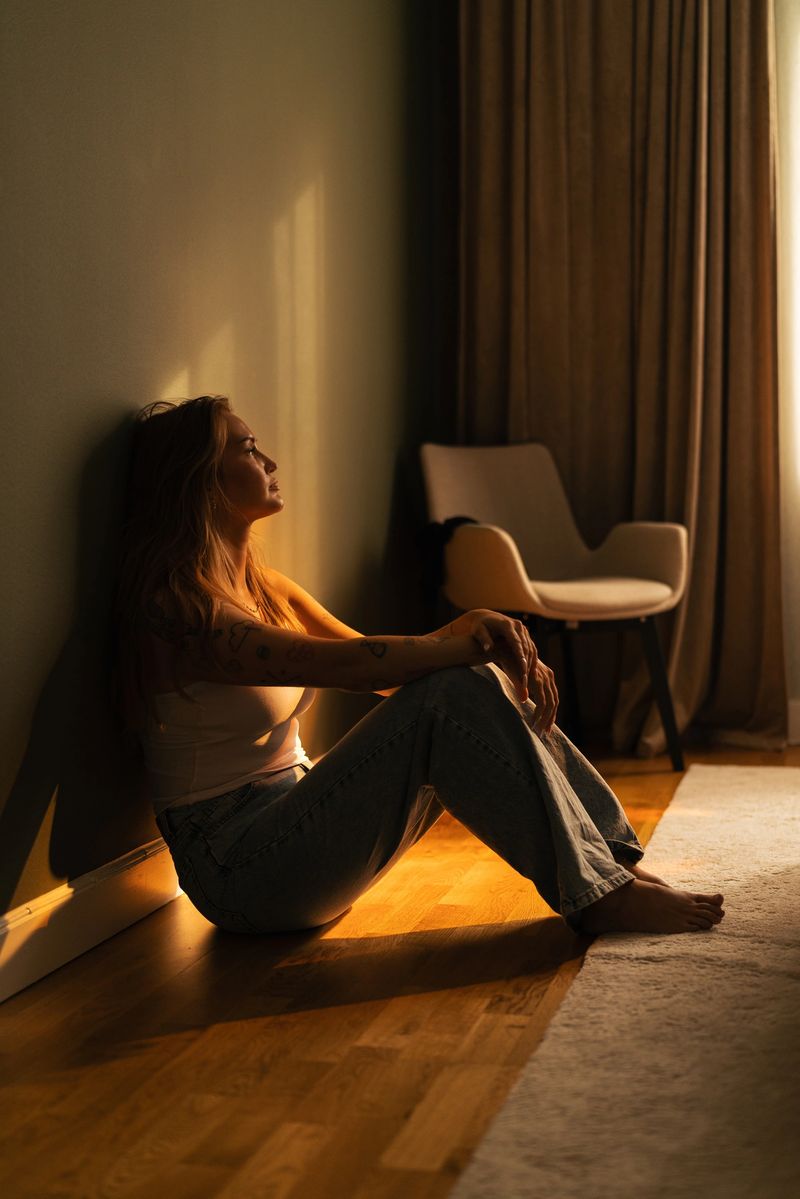Calm indoor capture of a woman sitting against a wall illuminated by golden sunlight, embracing peaceful introspection within a serene, modern living space featuring warm wood flooring and soft curtains.