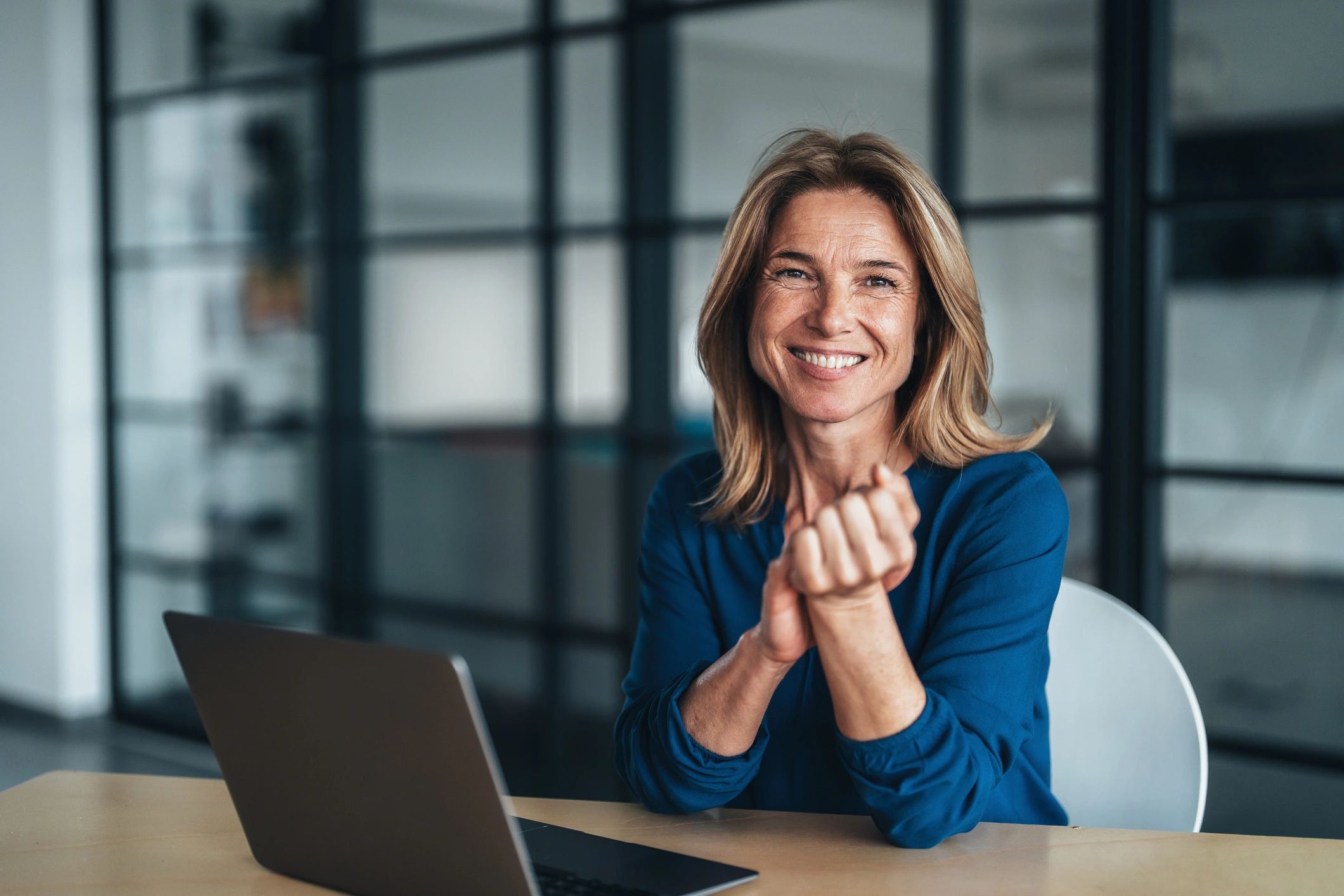 Smiling woman in blue shirt sitting with laptop in a modern office.