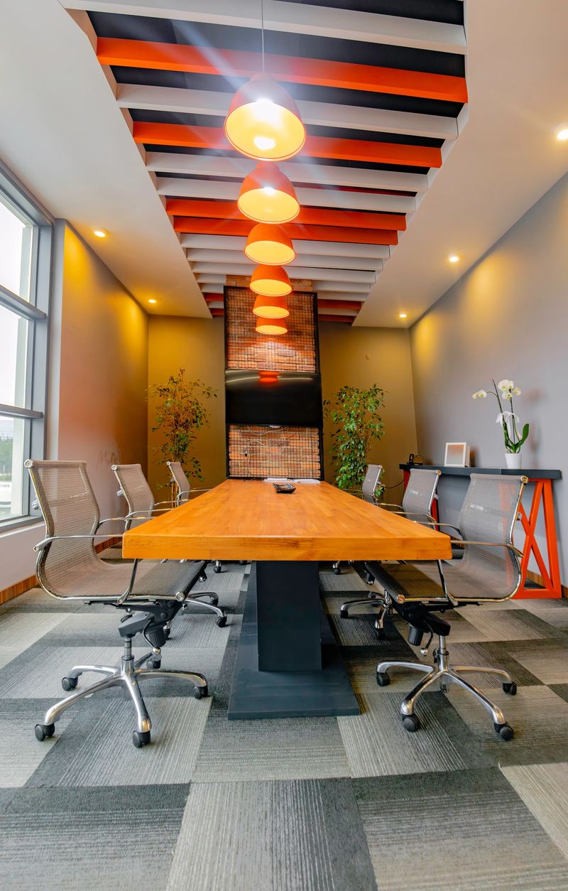 Interior view of a stylish and professional modern conference room featuring a long wooden meeting table, ergonomic mesh chairs on casters, and floor-to-ceiling windows allowing natural light to fill the space. The room’s clean lines, grey walls, orange ceiling lights, decorative plants, and minimalist decor create a sleek and inviting atmosphere ideal for board meetings, client presentations, and collaborative team sessions. The contemporary workspace reflects corporate innovation and architectural design suitable for creative agencies, tech companies, or executive environments