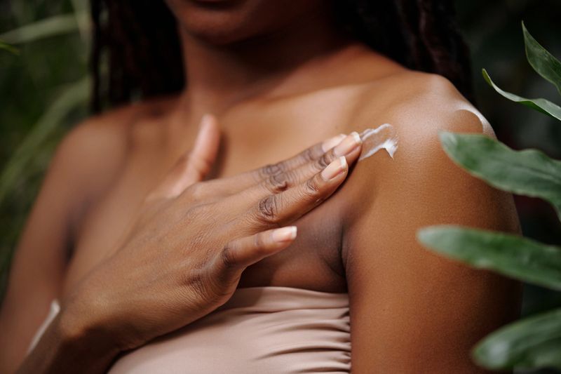 Close-up of girl applying moisturizing cream on shoulder with exposed skin surrounded by natural foliage and soft lighting creating serene and refreshing ambiance