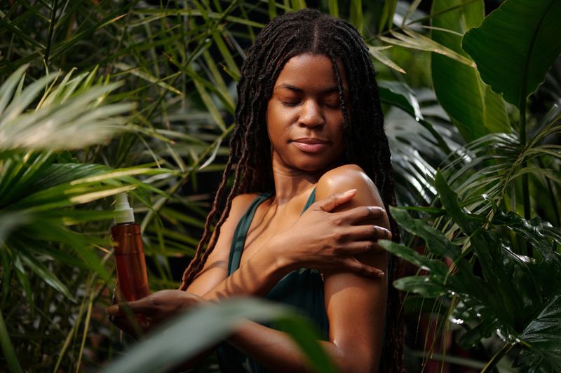 Black woman applying skincare product on her shoulder while standing amidst lush greenery. Relaxed expression and serene environment enhance self-care routine