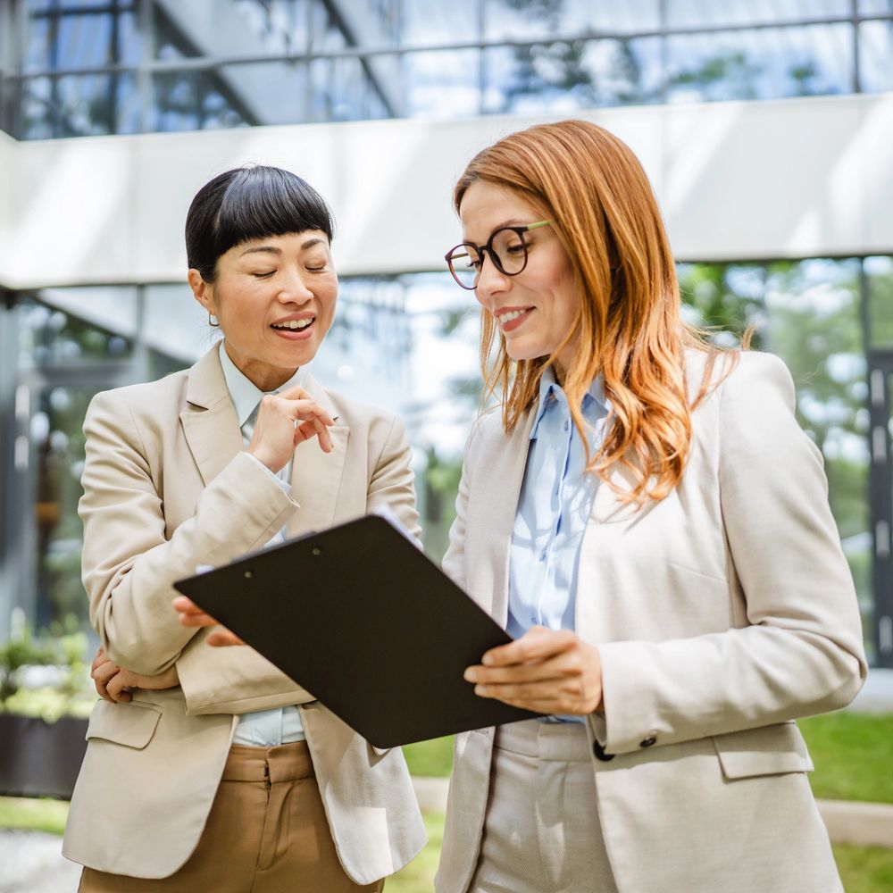 Two professional women discussing documents outdoors near an office building.