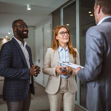 Three professionally dressed coworkers standing together in an office hallway, smiling and talking d