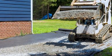 A skid steer is spreading gravel on a driveway near a house.