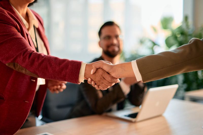 Two businesswomen shaking hands after successful meeting with their male colleague sitting in the background and looking at them, celebrating the agreement