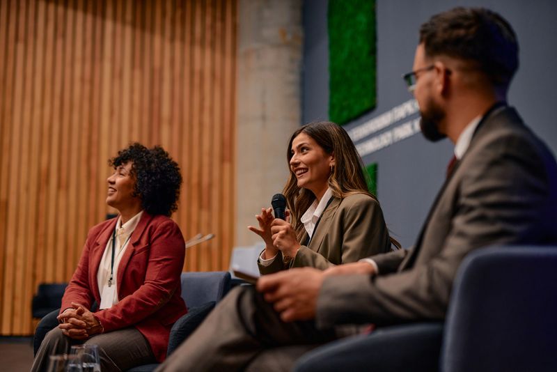 Businesswoman sitting at a conference. Speaking into a microphone while colleagues attentively listening to her insightful presentation. Fostering communication and collaboration among professionals