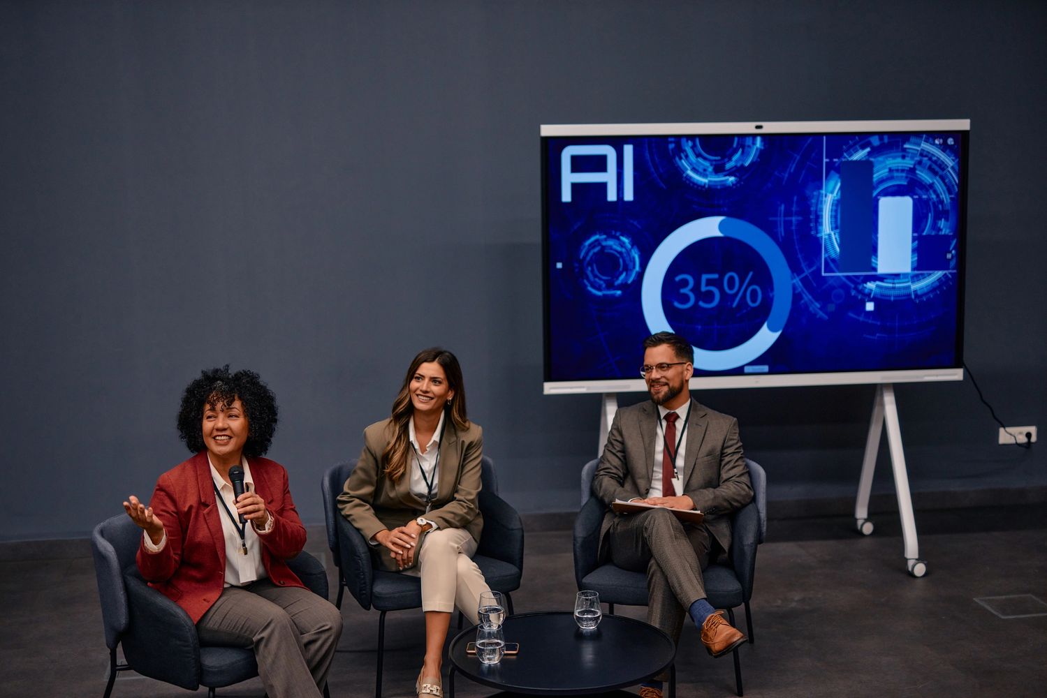 Three professionals discuss AI progress in a panel setting with a screen showing 35% completion.