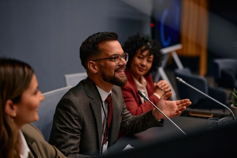 Group of politicians engaging in lively discussion at a press conference, with one member gesturing animatedly while smiling, creating an atmosphere of collaboration and communication
