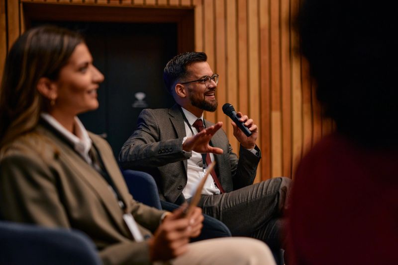 Businessman sitting and engaging with a microphone during a conference, sharing insights and ideas with the audience while fostering discussion and interaction among listeners