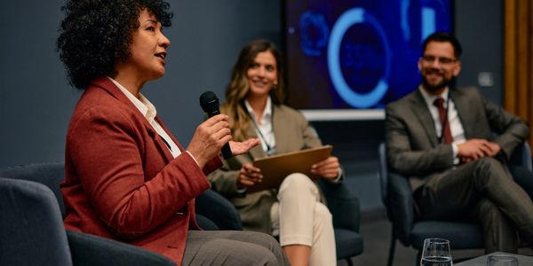 A woman speaking into a microphone during a panel discussion with two attentive colleagues.