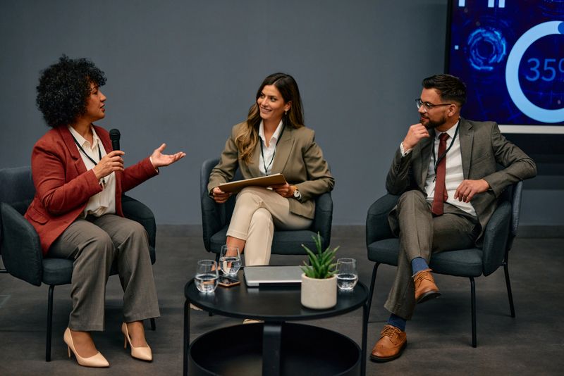 Three business professionals seated in armchairs engage in a lively discussion during a conference panel, with a woman actively speaking into a microphone, sharing insights and ideas