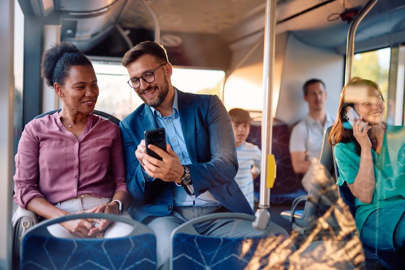 Two diverse coworkers are sitting together on the bus, sharing a funny moment on their smartphones, while other passengers are sitting behind them