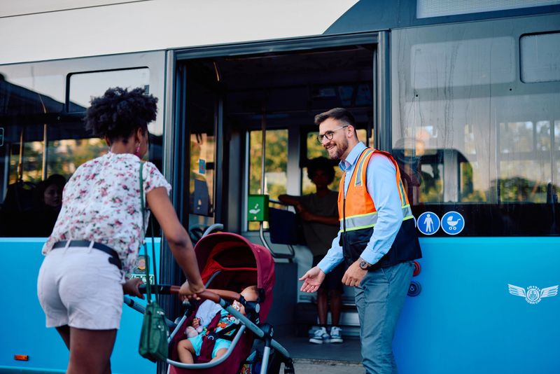 Friendly bus driver is assisting a young mother with a baby stroller to board a modern city bus, ensuring a smooth and accessible public transportation experience