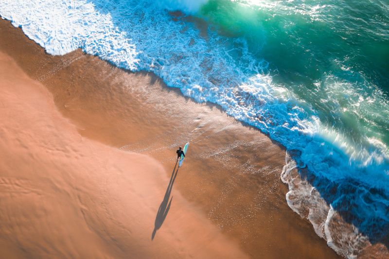 A lone surfer carrying a surfboard walks along the beach toward breaking waves, casting a long morning shadow on the sand. Shot from above, the image captures the peaceful anticipation of an early surf session.