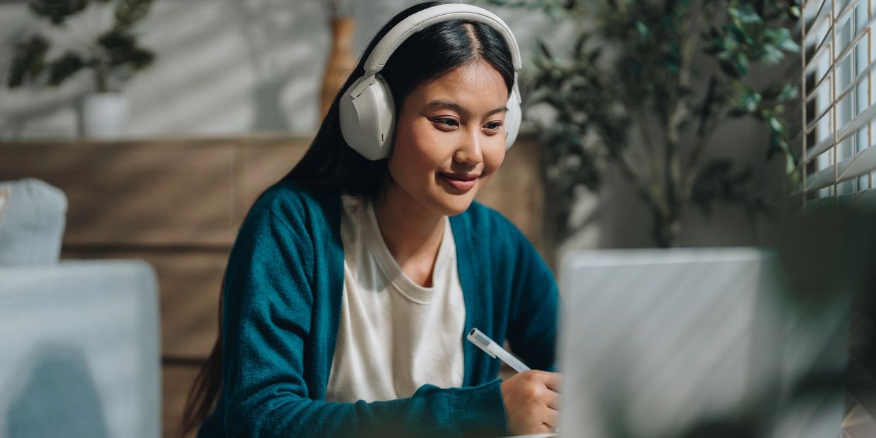 Teen student  with headphones sitting in front of a laptop taking notes.