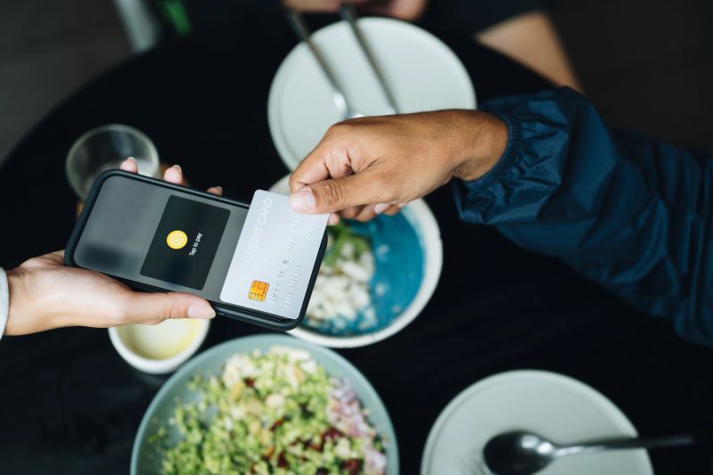 Close-up shot of man tapping a credit card on a smartphone to make a contactless payment at an organic healthy restaurant, capturing the seamless blend of modern technology and wellness culture, emphasizing the convenience of digital transactions in a clean, mindful dining environment