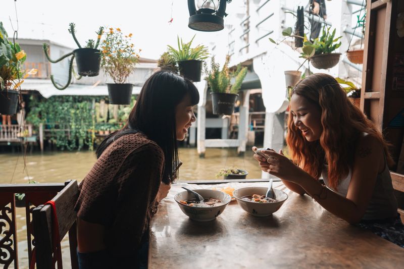 Two cheerful friends vlogging as they enjoy delicious local food, sharing their excitement with their followers, supporting local businesses, and savoring the rich culture of the area