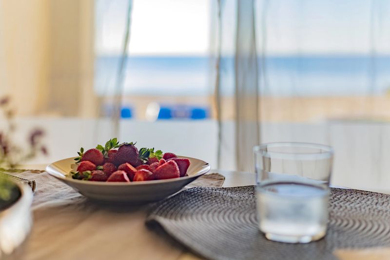 Altea. Alicante. Spain. A plate of fresh strawberries and a glass of water sit on a table with a beautiful beach view in the background, creating a serene and refreshing atmosphere