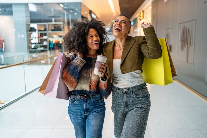 Two cheerful young women are walking through a mall, carrying shopping bags and a coffee cup, enjoying their successful shopping spree