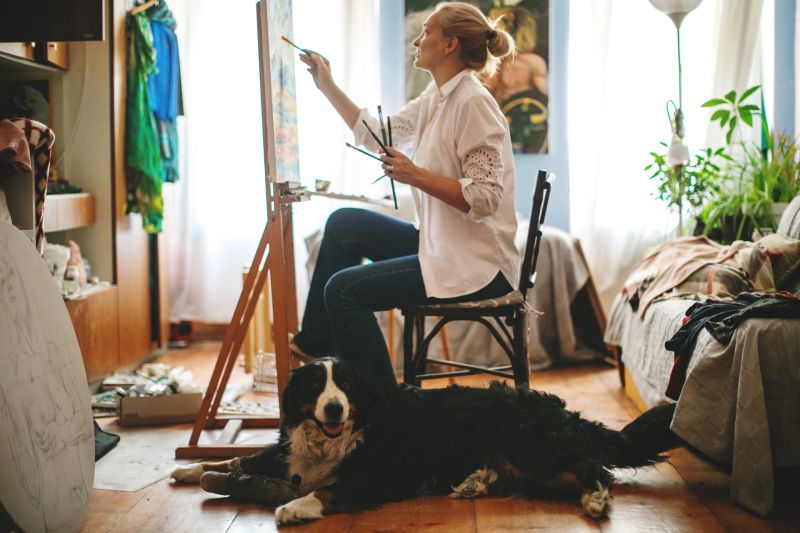A female artist painting on a canvas in her cozy home studio, while her Bernese Mountain Dog companion relaxes on the floor nearby. Natural ambient light and authentic domestic atmosphere. Candid moment of everyday artistic life.