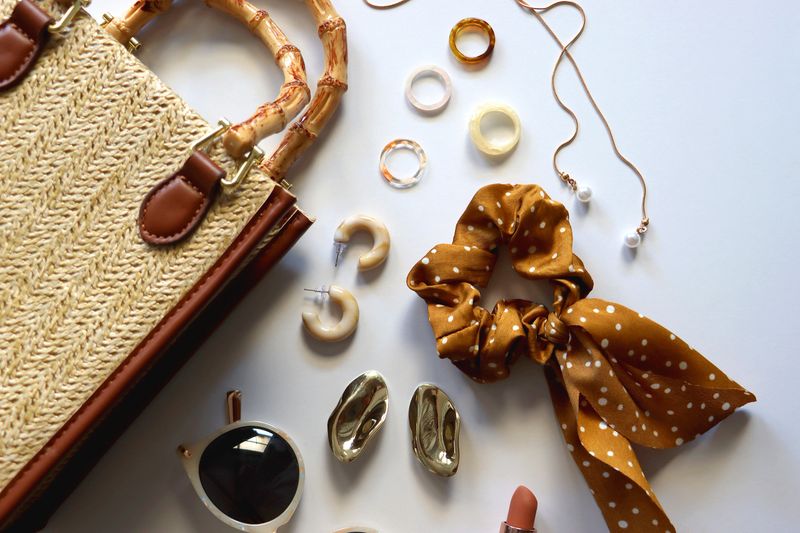 Wicker purse, sunglasses, scrunchie, lipstick and various gold and neutral jewelry on white background. Top view.