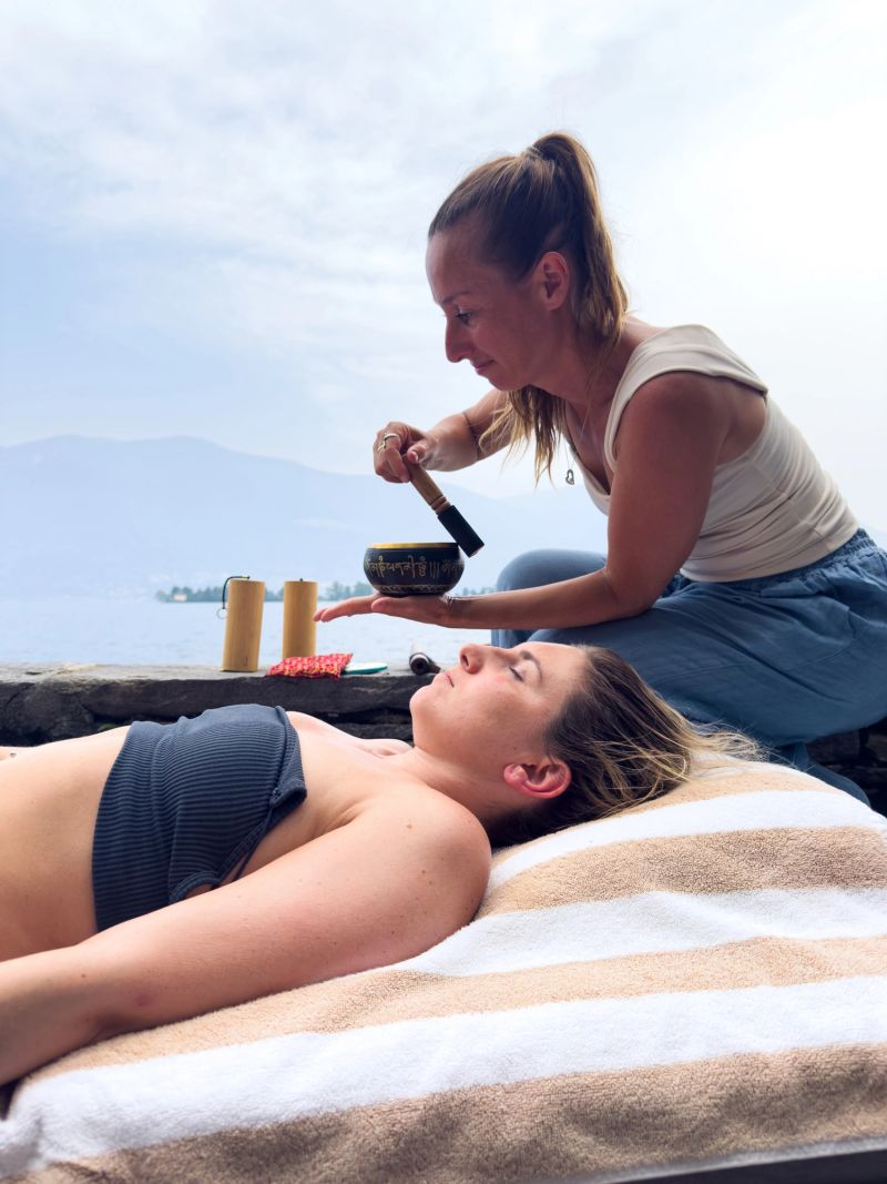 Practitioner using Tibetan singing bowl on woman's palm by lakeside. Outdoor alternative therapy session for neurodiverse individuals. Scenic relaxation and mindfulness retreat.