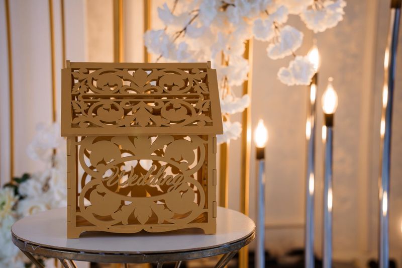 A beautiful laser-cut wooden card box adorned with "Wedding" script, illuminated amidst candles and cherry blossoms at a wedding reception, ideal for gift collection