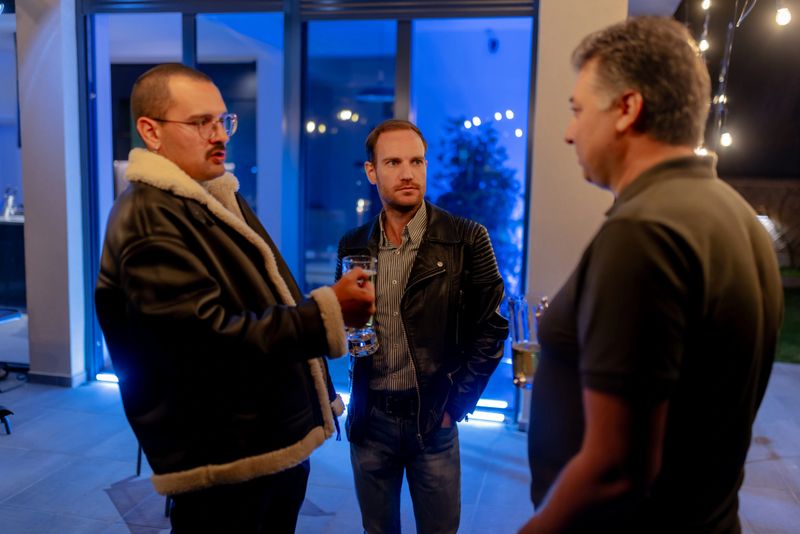 Three men interacting at a casual event, enjoying drinks and good company. The setting is modern, featuring aspects of outdoor and indoor ambiance accented with lighting.