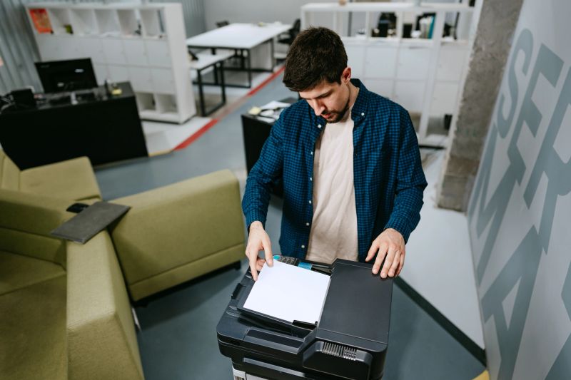 A man is preparing to scan documents at a multifunction printer in a contemporary office setting filled with modern furniture and ample light.