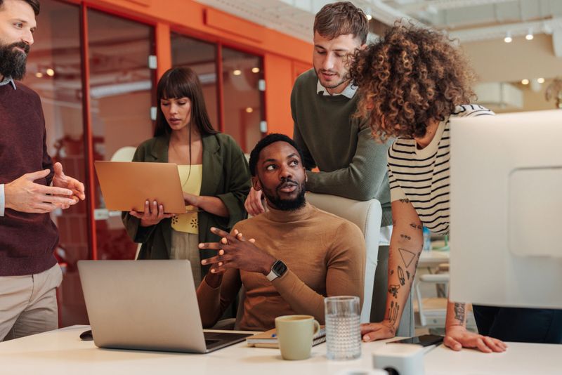 Group of young professionals are gathered around a desk, engaged in a collaborative discussion, using laptops and sharing their expertise to achieve a common goal
