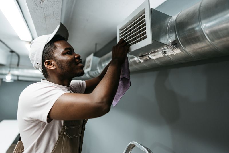 A person is engaged in cleaning an air vent in a modern industrial setting. Wearing a cap and gloves, they use a cloth to wipe the vent while standing on a ladder.